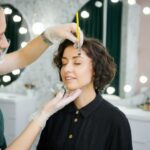 Happy woman keeping eyes closed and smiling while having eyebrow correction procedure in beauty salon. Female beauty specialist in sterile gloves shaping client eyebrows with cosmetic brow brush.