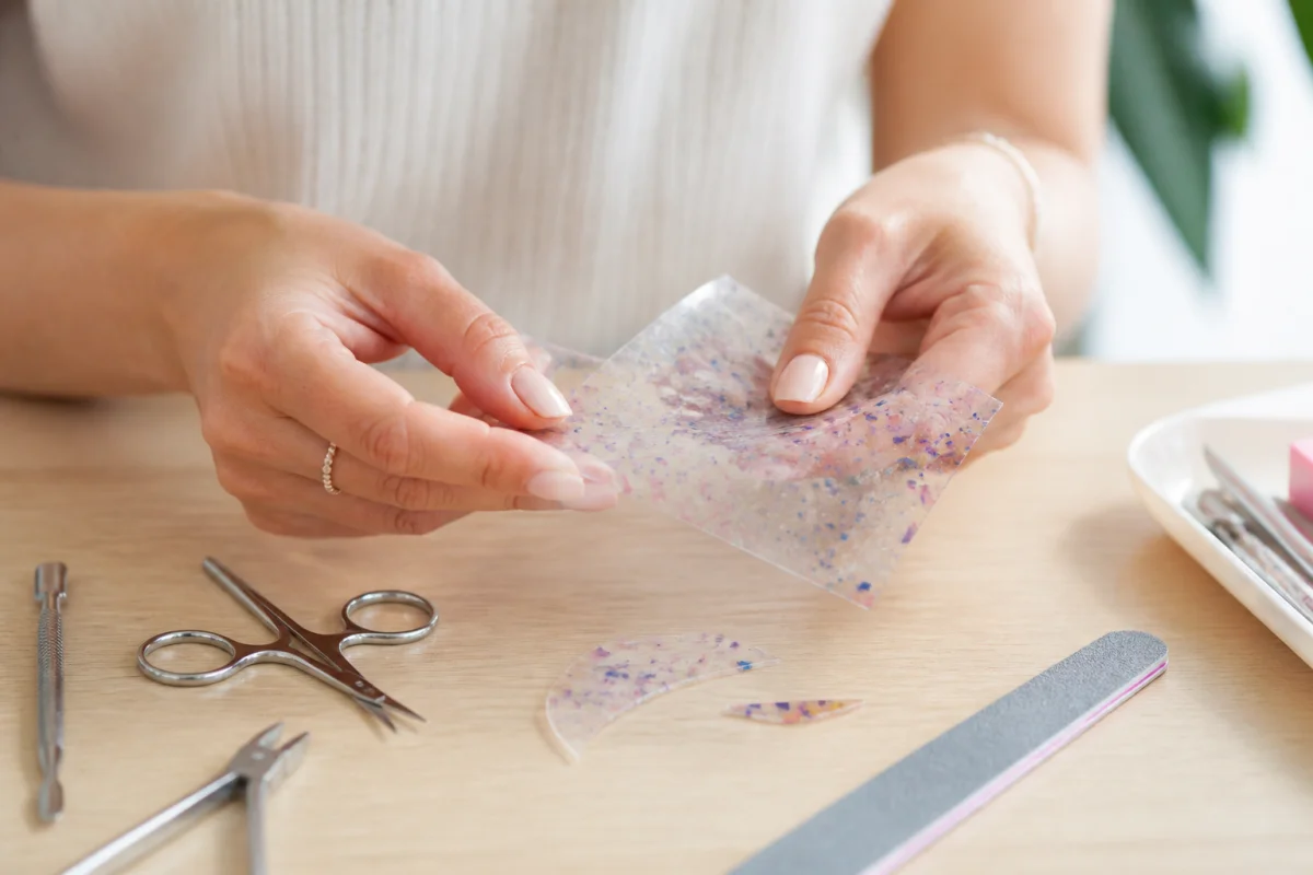 Mains découpant une plaque de plastique pour apprendre comment fabriquer des faux ongles personnalisés chez soi.